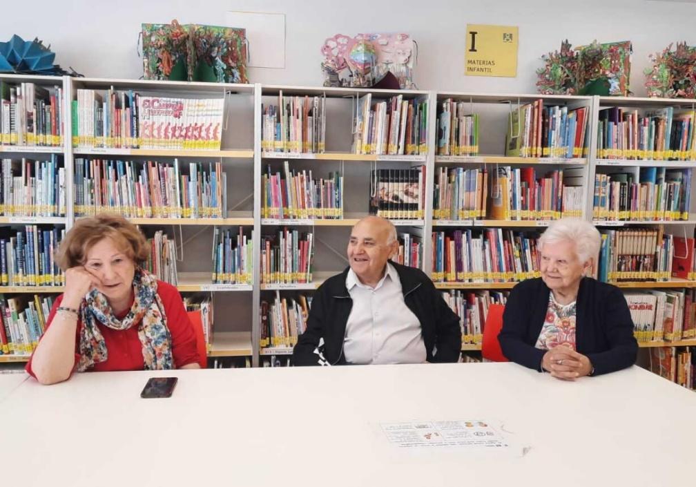 Salen 3 personas en una mesa de una biblioteca con libros de fondo. Se ve que salen sonriendo. Son personas del curso Mayores Conectados