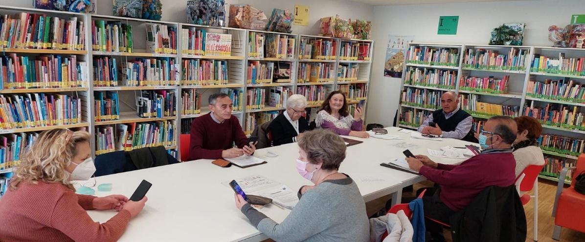 Salen unas 7 personas en una mesa de una biblioteca con libros de fondo. Salen mirando al móvil. Se nota que están aprendiendo a utilizar el móvil. Se ve que salen sonriendo. Son personas del curso Mayores Conectados